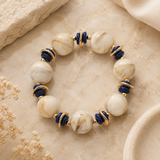 A beaded stretch bracelet featuring large white and cream marbled round beads, deep blue cylindrical beads, and small gold-toned spacer rings, displayed on a textured beige surface.