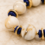 A close-up of a beaded strand featuring large white and cream marbled glass beads, accented with dark blue discs and textured gold spacer rings, resting on a light beige stone surface.