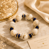 A beaded stretch bracelet with large cream and white marbled beads, deep blue beads, and gold spacer rings, styled on a beige stone slab with a cream fabric and dried flowers.