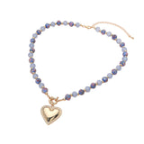 A gold-tone beaded necklace with light blue and purple iridescent beads, featuring a dangling gold heart-shaped pendant with a textured border, shown on a plain white background.