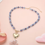 A beaded necklace with light blue and purple iridescent beads, gold spacer rings, and a gold heart-shaped pendant, displayed on a soft pink background with decorative heart props.