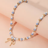 Blue floral ceramic bead necklace featuring pearls, gold accents, and a delicate gold bow pendant on a soft pink background