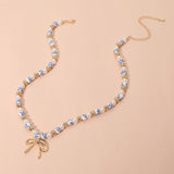 Blue floral ceramic bead necklace featuring pearls, gold accents, and a delicate gold bow pendant on a soft pink background