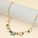 Gold charm station necklace featuring a colorful enamel butterfly, pearl, heart, flower, bow and cross charms with decorative black bead accents on alight background