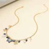 Gold charm station necklace featuring a colorful enamel butterfly, pearl, heart, flower, bow and cross charms with decorative black bead accents on beige background 
