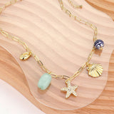 Coastal gold paperclip chain necklace with starfish, shell and natural stone charms on a wooden surface.