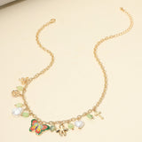 A gold chain necklace with multiple charms, including a colorful butterfly, a white pearl, a green stone, a small flower, and a cross, displayed on a light beige background.