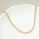 A gold tennis necklace paved with clear round-cut stones, displayed on a white curved jewelry stand against a light gray background.
