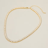 A gold tennis-style necklace with clear rhinestones, shown on a plain light beige background. The necklace has a lobster clasp and an adjustable chain extender.