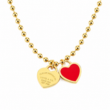 Gold ball chain necklace with two heart-shaped charms, one gold-toned engraved and one red enamel, on a white background.