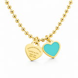 A gold ball chain necklace with two heart-shaped tags, one gold-toned engraved tag and one with a turquoise blue enamel center, shown on a white background.