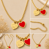 A collage showing multiple views of a gold-tone ball-chain necklace with two heart pendants: one polished gold heart engraved with 'PLEASE RETURN LOVE & CARE' and one red heart pendant. The collage is set on a warm beige background with decorative dried flowers and satin.