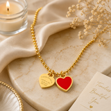 Gold ball-chain necklace with two heart charms, one engraved gold and one solid red, styled on a beige marble slab with a lit candle, satin fabric, dried flowers, and a card.