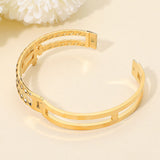 Gold stainless steel double row hinged bangle bracelet featuring sculptural interlock accents and polished finish on a beige background. 