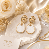 Gold geometric drop earrings featuring polished organic gold-tone studs with white marbled resin dangle accents on a beige background.