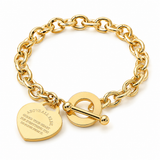 A gold-tone chain bracelet with a toggle clasp and two charms: a heart-shaped engraved charm and a round ring charm, shown on a plain white background.