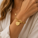 A close-up of a woman's wrist wearing a gold chain bracelet with a heart-shaped engraved charm and a toggle clasp, styled with a white blouse.