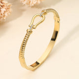 Gold stainless steel twisted hinged bangle bracelet with horsebit buckle accent and crystal details on a beige background