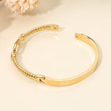 Gold stainless steel twisted hinged bangle bracelet with horsebit buckle accent and crystal details on a beige background