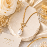 A gold chain necklace with a toggle clasp and a teardrop-shaped white pearl pendant, styled on a textured cream stone slab with a cream rose, dried flowers, a perfume bottle, and a card reading 'DAZZLED BY JEWELS'.