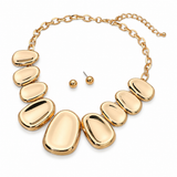 Gold statement necklace with seven smooth, polished oval stone-like discs connected by a gold chain, shown with matching stud earrings on a white background.