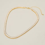 A gold tennis-style choker necklace with clear rectangular gemstones, shown on a plain light beige background. The necklace is arranged in an oval shape with a small clasp and extender chain visible at the top right.