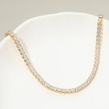 A gold tennis-style necklace with clear square-cut stones, displayed on a white curved surface against a light gray background.