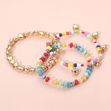 Three-piece colorful stretch bracelet set featuring multicolored beads, gold-tone heart charms, and layered stackable designs on a pink background