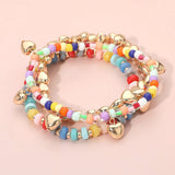Three-piece colorful stretch bracelet set featuring multicolored beads, gold-tone heart charms, and layered stackable designs on a pink background