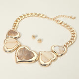 Gold heart-link statement necklace with matching stud earrings, featuring polished metallic gold heart-shaped links with iridescent beige inlay stones, displayed on a light cream background.