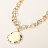 Gold chunky chain necklace with organic round plate pendant and modern statement design on a light beige background