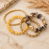 Three beaded stretch bracelets arranged on a light marble surface with dried grass and a stone prop. The bracelets feature gold-tone beads, translucent yellow beads, and irregular agate-like stones in gray, brown, and white.