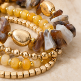 A close-up of a multi-strand beaded bracelet stack featuring gold-tone metal beads, yellow and amber stone beads, and natural agate chips in brown, cream, and white on a light beige surface.