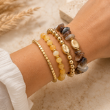 A close-up of a person's wrist wearing four stacked beaded bracelets: a gold chain bracelet, a gold bead bracelet, a yellow stone bracelet, and a mixed-color stone bracelet with gold-toned accents.