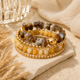 A stack of four beaded stretch bracelets displayed on a light stone slab. The bracelets feature a mix of gold-plated beads, translucent yellow and white beads, and natural brown, gray, and gold stone beads.
