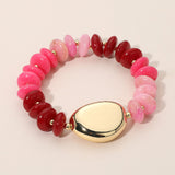 A beaded stretch bracelet featuring polished pink and deep red round beads, separated by small gold spacer beads, with a large smooth cream-colored oval centerpiece and gold end caps, shown on a light beige background.
