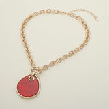 A gold chain necklace featuring a large, organic-shaped red pendant with a gold border, displayed on a light beige background.