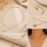 A collage showing a gold double-layered snake chain necklace with a small heart pendant, displayed on neutral fabric and draped with dried flowers, with one panel showing the necklace worn on a model's neck.