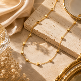 A delicate gold chain necklace with small cube-shaped beads draped over a textured beige stone slab, styled with cream satin fabric, dried flowers, and a gold-rimmed dish.