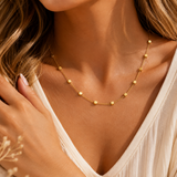 A close-up lifestyle shot of a model wearing a delicate gold chain necklace with small, evenly spaced cube-shaped beads. The model is wearing a white V-neck top, and her hand rests near her shoulder.