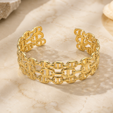 A gold-toned open cuff bracelet with an interlocking link pattern, displayed on a light beige marble surface.