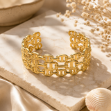 A wide gold cuff bracelet with an interlocking link pattern, displayed on a white marble slab with dried flowers and a seashell in a warm, sunlit setting.