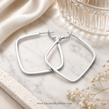 A pair of silver-toned open hoop earrings with a rounded square design, displayed on a white marble surface with cream satin fabric, a glass bowl, and small dried flowers.