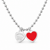 A silver ball chain necklace with two heart-shaped charms, one polished silver heart engraved with 'PLEASE RETURN TO LOVE & CO. SINCE 1947 NEW YORK' and one red heart charm, shown on a white background.