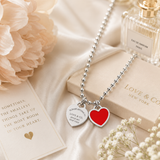 A silver ball-chain necklace with two heart charms, one red and one engraved silver, styled on a beige satin fabric with flowers, a perfume bottle, and a card.