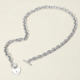 A silver chain bracelet with a heart-shaped charm and a toggle clasp, displayed on a light beige background.