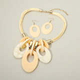 A multi-strand statement necklace with matching oval drop earrings, featuring cream, beige, and white textured disc elements with gold-tone metal hardware, displayed on a neutral gray background.