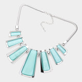 A statement bib necklace featuring multiple angular light aqua blue stone inlays set in silver-tone metal, suspended from a silver snake chain, shown on a light gray background.